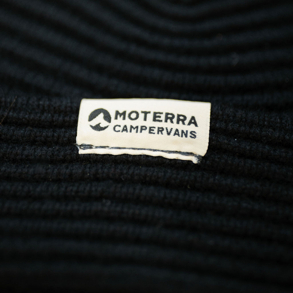 Close-up of a white woven patch with the Moterra Campervans logo, stitched onto a ribbed black beanie.