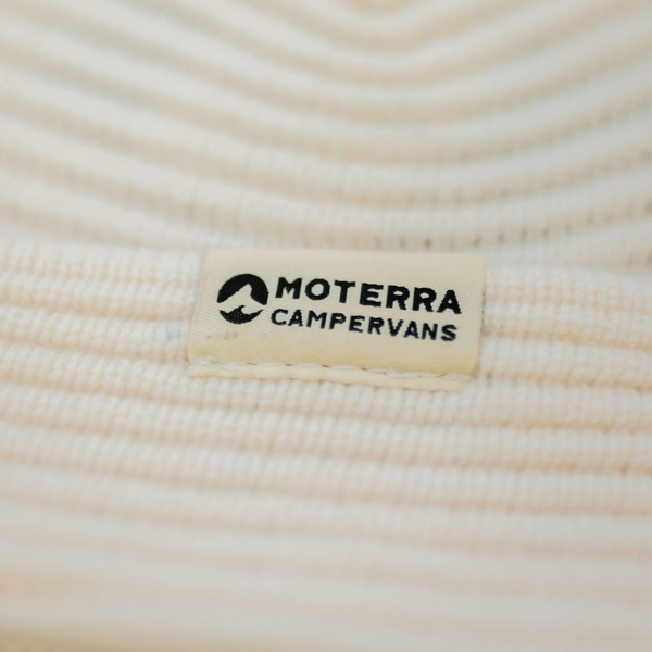 Close-up of a white woven patch with the Moterra Campervans logo, stitched onto a ribbed cream beanie.
