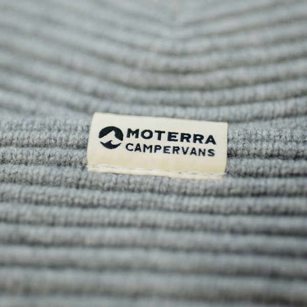 Close-up of a white woven patch with the Moterra Campervans logo, stitched onto a ribbed grey beanie.