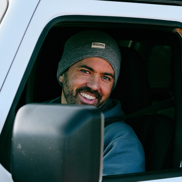 Smiling man in a Moterra Campervan wearing a gray ribbed beanie with a Moterra Campervans logo patch.